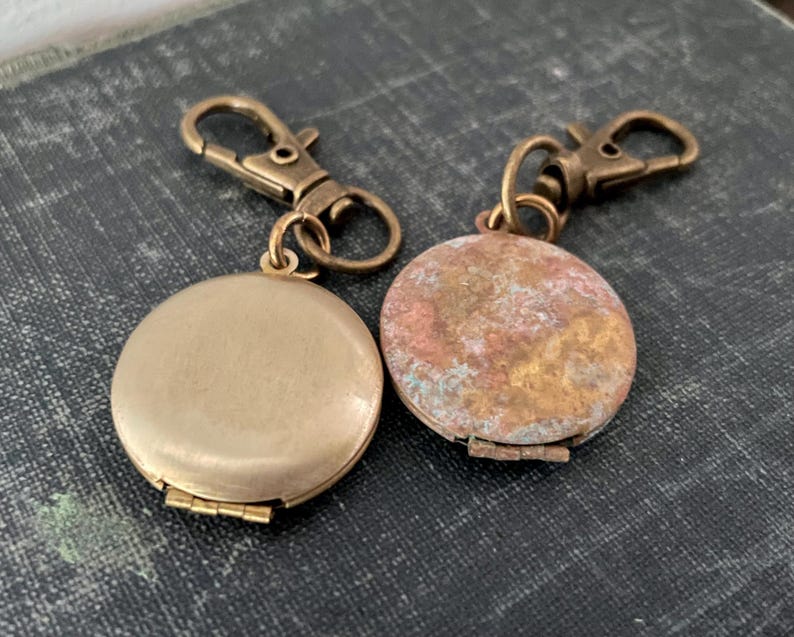 May include: Two gold-toned locket keychains with a clasp closure. One locket has a smooth, polished finish, while the other has a textured, aged finish.