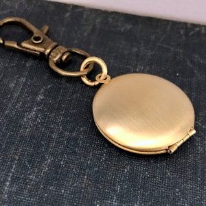 May include: A gold-toned locket keychain with a brushed finish. The locket has a hinged lid and a clasp for attaching to a key ring.