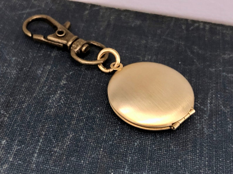 May include: A gold-toned locket keychain with a brushed finish. The locket has a hinged lid and a clasp for attaching to a key ring.