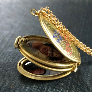 May include: A gold-toned triple locket necklace with a chain. The locket is open, revealing three oval-shaped photo compartments. The locket is decorated with floral designs in red, blue, and green.