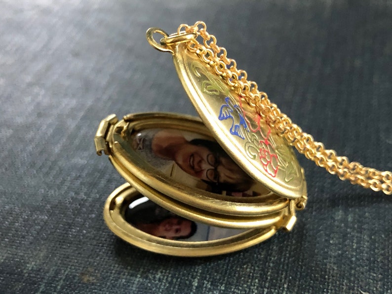 May include: A gold-toned triple locket necklace with a chain. The locket is open, revealing three oval-shaped photo compartments. The locket is decorated with floral designs in red, blue, and green.