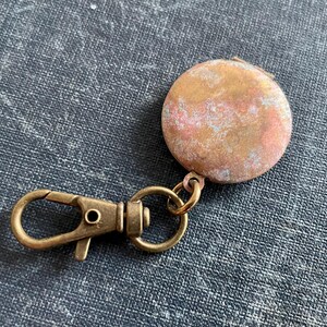 May include: A round, brass locket with a worn, aged finish. The locket is attached to a brass key chain with a spring clasp.