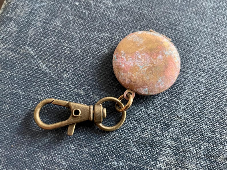 May include: A round, brass locket with a worn, aged finish. The locket is attached to a brass key chain with a spring clasp.