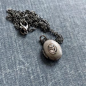 May include: An oval-shaped, silver-toned locket necklace with a detailed rose design on the front. The locket hangs from a matching chain, resting on a textured blue surface. The chain is a dark silver color.