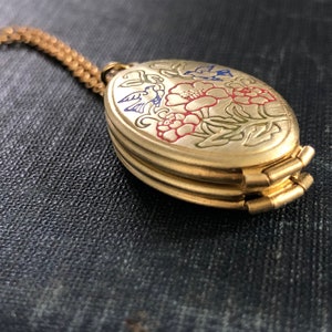 May include: A gold-tone oval locket necklace with a floral design featuring red, blue, and green enamel. The locket has a hinged opening and is attached to a gold-tone chain.