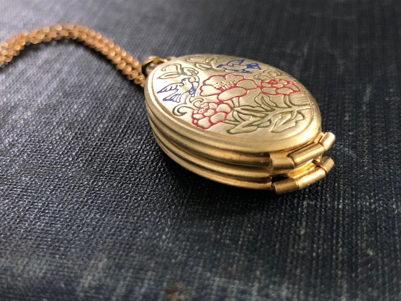 May include: A gold-tone oval locket necklace with a floral design featuring red, blue, and green enamel. The locket has a hinged opening and is attached to a gold-tone chain.