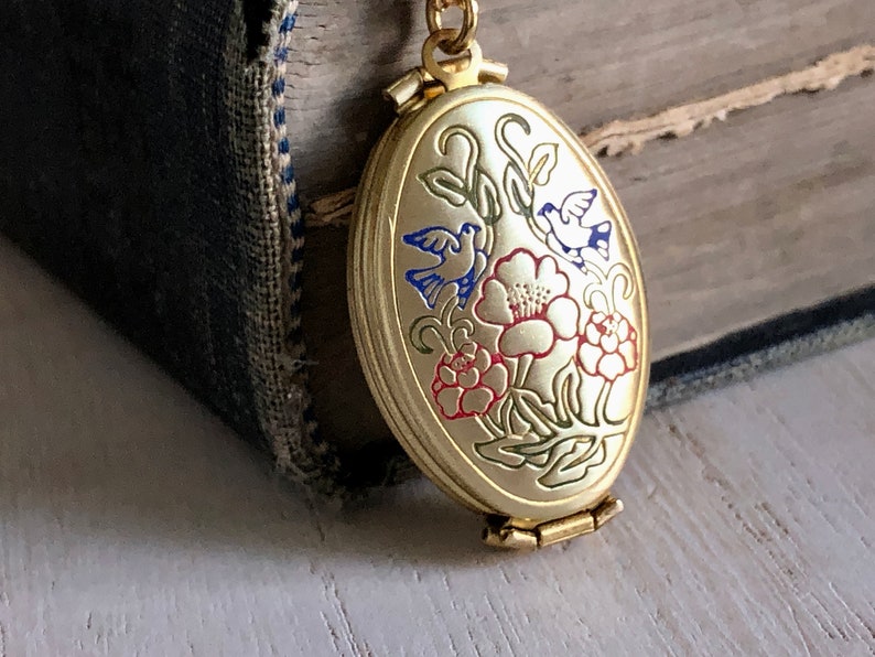 May include: Gold oval locket with a hinged closure. The locket features an enamel design of two blue birds and red flowers on a green stem.