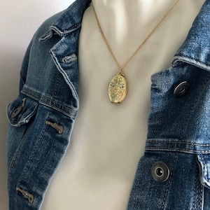 May include: A gold chain necklace with a gold oval locket pendant featuring a floral design. The necklace is worn by a person wearing a white shirt and a blue denim jacket.