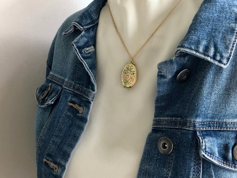 May include: A gold chain necklace with a gold oval locket pendant featuring a floral design. The necklace is worn by a person wearing a white shirt and a blue denim jacket.