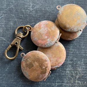 May include: A set of five small, round, brass-colored locket charms with a worn, distressed finish. The charms are attached to a brass-colored key ring.