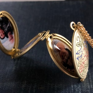 May include: A gold-toned oval locket with a chain. The locket has two compartments, each with a photo of a person inside. The locket is decorated with a floral design in red, blue, and green.
