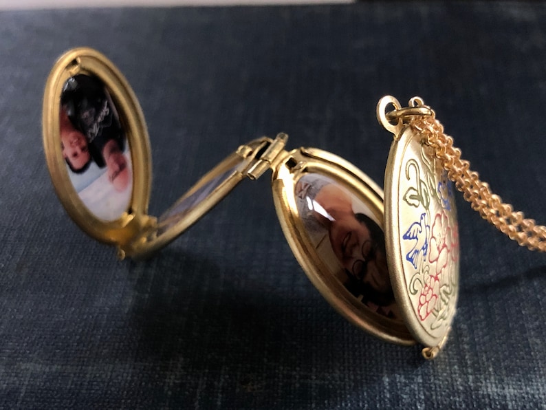 May include: A gold-toned oval locket with a chain. The locket has two compartments, each with a photo of a person inside. The locket is decorated with a floral design in red, blue, and green.
