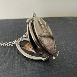 May include: Silver locket necklace with a chain. The locket is open and shows a photo of a cat inside. The locket has an engraved design of a bird on the outside.