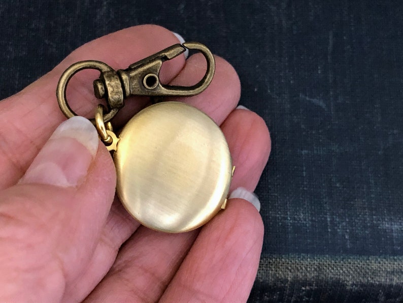 May include: A gold-toned locket with a round, smooth surface. The locket is attached to a gold-toned chain with a clasp.