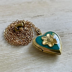 May include: A gold-coloured heart-shaped locket with a teal enamel background and a gold flower design. The locket is on a gold chain. The jewellery is displayed on a light-coloured wooden surface.