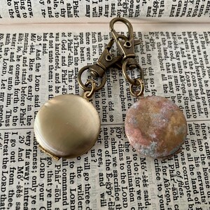 May include: A gold-toned keychain with two round, decorative charms. One charm is a smooth, light gold color, while the other is a textured, brown and white color.