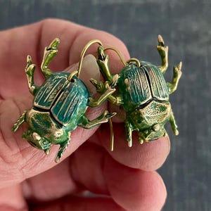Puede incluir: Un par de pendientes con forma de escarabajo. Los pendientes presentan una combinación de colores verde azulado y verde con detalles dorados. El cuerpo del escarabajo es verde azulado, y las patas y antenas son verdes.