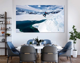 May include: A snowboarder jumps off a snow-covered cliff in Antarctica. The sky is blue with white clouds. The water is a deep blue. The snow is white and glistening.