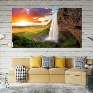 May include: A scenic landscape photograph featuring a waterfall cascading down a rocky cliff face. The sky is a vibrant orange and pink, with fluffy white clouds. Lush green vegetation surrounds the waterfall.