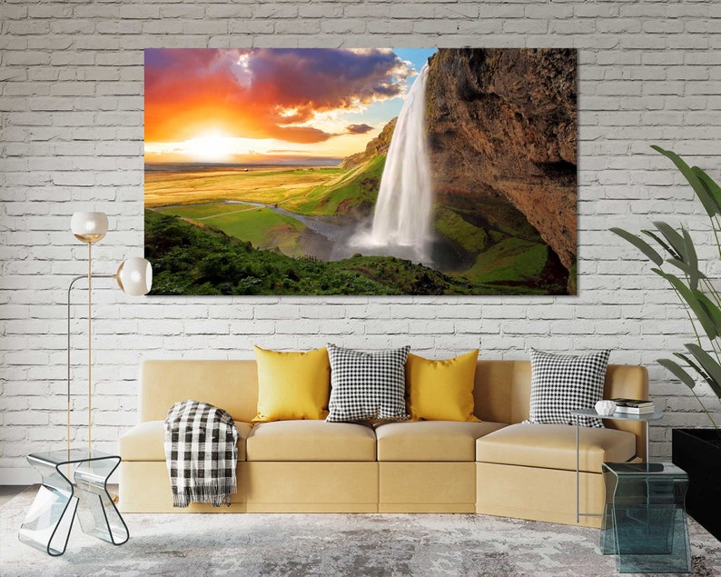 May include: A scenic landscape photograph featuring a waterfall cascading down a rocky cliff face. The sky is a vibrant orange and pink, with fluffy white clouds. Lush green vegetation surrounds the waterfall.