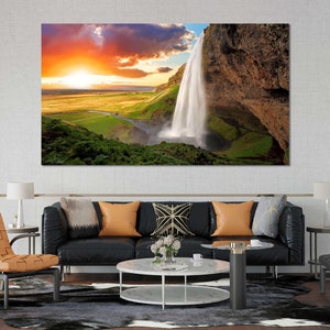 May include: A scenic landscape photograph featuring a waterfall cascading down a rocky cliff face. The sky is a vibrant orange and pink, with clouds reflecting the sunset. Lush green vegetation surrounds the waterfall.