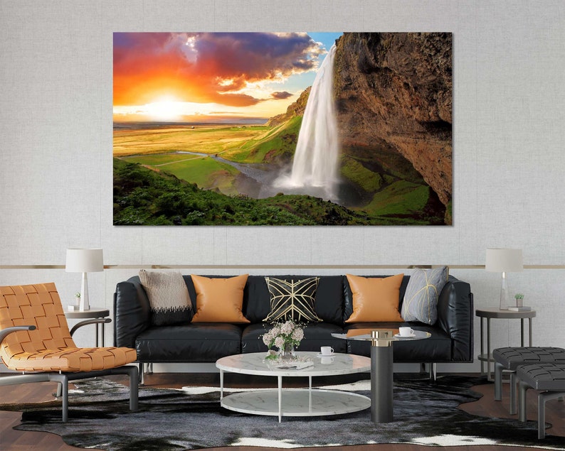May include: A scenic landscape photograph featuring a waterfall cascading down a rocky cliff face. The sky is a vibrant orange and pink, with clouds reflecting the sunset. Lush green vegetation surrounds the waterfall.