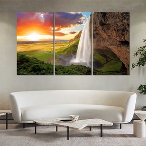 May include: A three-panel canvas print featuring a scenic landscape with a waterfall cascading down a rocky cliff. The sky is a vibrant orange and pink, with clouds in the background. The waterfall is surrounded by lush green vegetation.