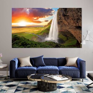 May include: A scenic landscape photograph featuring a waterfall cascading down a rocky cliff face. The sky is a vibrant orange and pink, with fluffy white clouds. Lush green vegetation surrounds the waterfall.