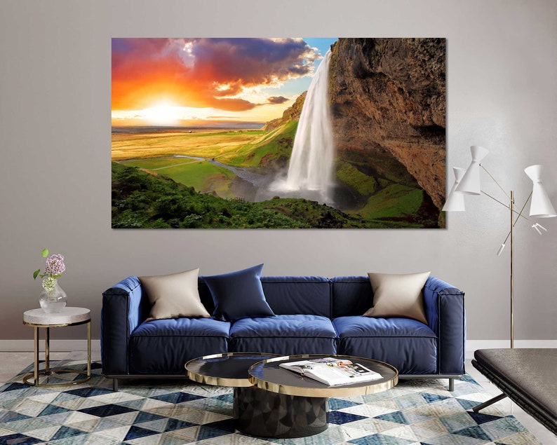 May include: A scenic landscape photograph featuring a waterfall cascading down a rocky cliff face. The sky is a vibrant orange and pink, with fluffy white clouds. Lush green vegetation surrounds the waterfall.