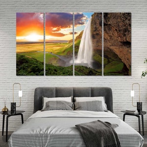 May include: A multi-panel canvas print featuring a scenic landscape with a waterfall cascading down a rocky cliff. The sky is a vibrant orange and yellow, with clouds in the background. The scene is framed by lush green vegetation.