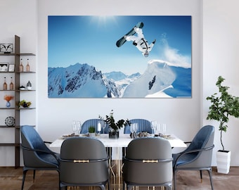 May include: A snowboarder performs a trick in mid-air, with a snowy mountain range in the background. The sky is blue and sunny.