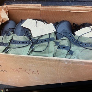 May include: A cardboard box filled with several pairs of green and black military boots. The boots have black laces and eyelets, and are partially wrapped in brown paper. The box has handwritten numbers and text on the side.