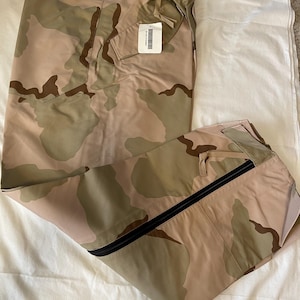 以下が含まれることがあります： 側面に黒いジッパーが付いた迷彩柄のパンツ。パンツは半分に折り畳まれ、ウエストにタグが付いています。