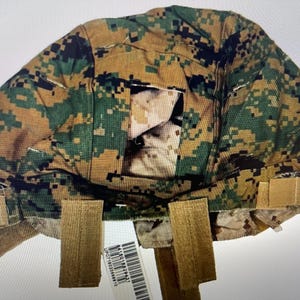 Puede incluir: Una capucha con estampado de camuflaje con múltiples correas de velcro color beige. La capucha presenta un patrón de camuflaje digital en tonos verdes, marrones y beige. La capucha está diseñada para usarse sobre la cabeza y los hombros.
