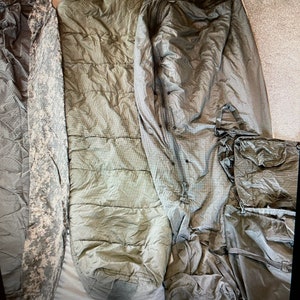 May include: Three sleeping bags, one in a camouflage pattern, one in a green color, and one in a dark gray color. The sleeping bags are laid out on a white surface.