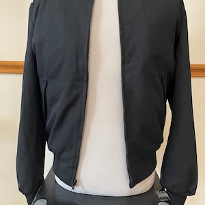 Pode incluir: Um blusão bomber preto sólido com fecho de correr frontal a todo o comprimento, gola subida e punhos canelados nas mangas e na bainha. O blusão tem mangas compridas e dois bolsos laterais, apresentado num manequim para mostrar a estrutura.