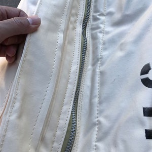 May include: Close-up of a white canvas bag with a zipper and stitching detail. The zipper is silver and the stitching is a light green color.