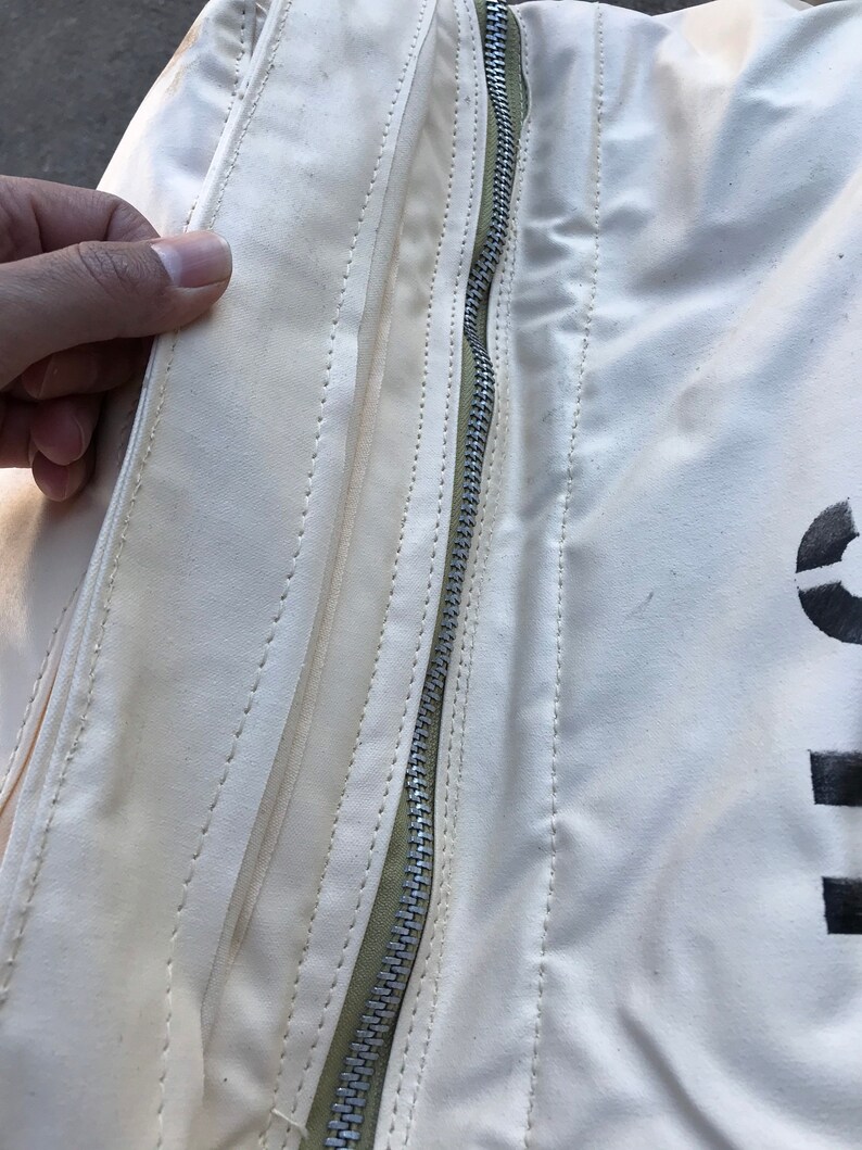 May include: Close-up of a white canvas bag with a zipper and stitching detail. The zipper is silver and the stitching is a light green color.