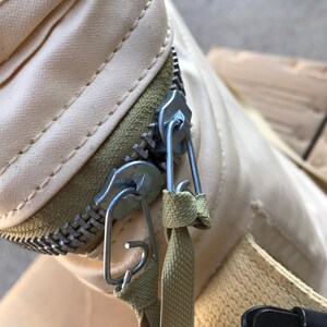 May include: Close-up of a cream-colored zipper with a metal slider and two metal clips. The zipper is attached to a beige fabric strap.