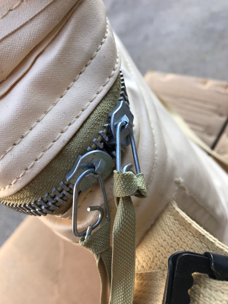 May include: Close-up of a cream-colored zipper with a metal slider and two metal clips. The zipper is attached to a beige fabric strap.