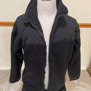 May include: A black fleece jacket with a full zipper closure. The jacket features a high collar and reinforced panels on the shoulders and elbows. The jacket is displayed on a mannequin.