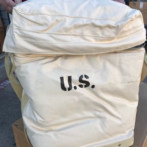 May include: A large, white canvas bag with a zippered top and a black "U.S." printed on the front. The bag has a brown leather strap and a brown leather handle.