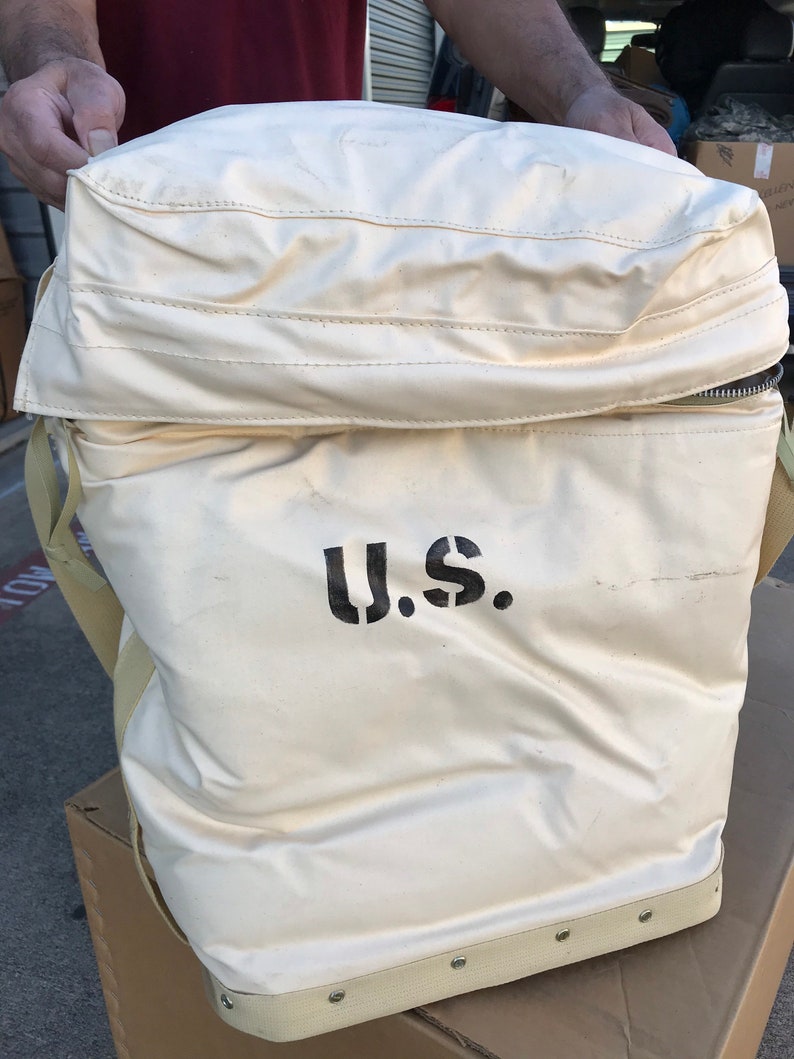 May include: A large, white canvas bag with a zippered top and a black "U.S." printed on the front. The bag has a brown leather strap and a brown leather handle.