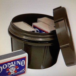以下が含まれることがあります： 暗いオリーブグリーンの容器と、部分的に開いたお揃いの蓋。Domino Diamond Matchesの箱がいくつか見えます。前景のマッチ箱には、Dominoのロゴとテキストが表示されています。