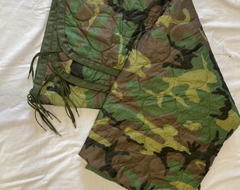 May include: A quilted liner featuring a woodland camouflage pattern in shades of green, brown, and black. The garment has a solid olive green trim along the edges and includes thin olive green fabric ties for fastening at the side.