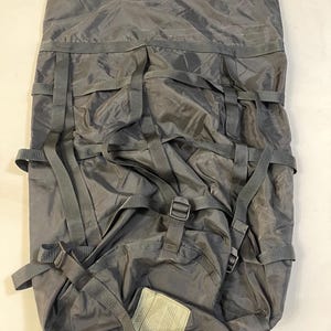 May include: A grey nylon backpack with multiple straps and buckles. The bag has a drawstring closure at the top and a small label attached near the bottom. The material appears to be water-resistant.