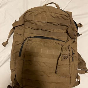 Könnte beinhalten: Ein beigefarbener taktischer Rucksack mit mehreren Fächern und Riemen. Der Rucksack verfügt über ein MOLLE-System zur Befestigung zusätzlicher Ausrüstung, einen oberen Griff und verstellbare Schultergurte. Die Reißverschlüsse sind schwarz.