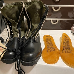May include: Black and olive green military boots with laces and a tag. The boots have a leather lower and canvas upper. Two tan insoles with text are next to the boots. The tag reads "BOOTS, HOT WEATHER".