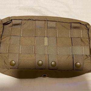May include: A brown tactical pouch with a zipper closure and multiple webbing straps. The pouch has four brown plastic buttons on the front.