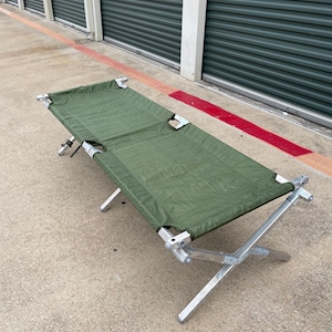 May include: A green canvas cot with a silver metal frame. The cot is folded up and sitting on a concrete surface.
